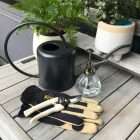 Gardening tools on a wooden table: a black watering can, a glass plant mister, a pair of beige and black gloves, and a small trowel, with potted ferns and succulents in the background. - Quality Garden Supplies