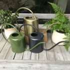 Several metal watering cans in green, dark gray, and brass sit on a wooden deck among potted plants. - Quality Garden Supplies