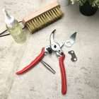 ryset-replacement-compact-blade-qualitygardensupplies-03 - Quality Garden Supplies Red-handled pruning shears with spare blades and a small metal plate, laid on a light tile floor beside a spray bottle and a broom. - Quality Garden Supplies